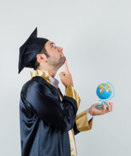 Young man holding school globe in graduate uniform and looking pensive .