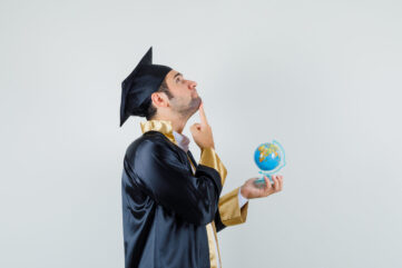 Young man holding school globe in graduate uniform and looking pensive .